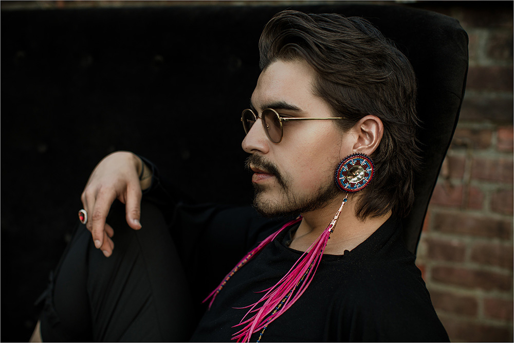 The author Joshua Whitehead sits in side profile, dressed in black, wearing sunglasses, a pinky ring, and pink earrings.
