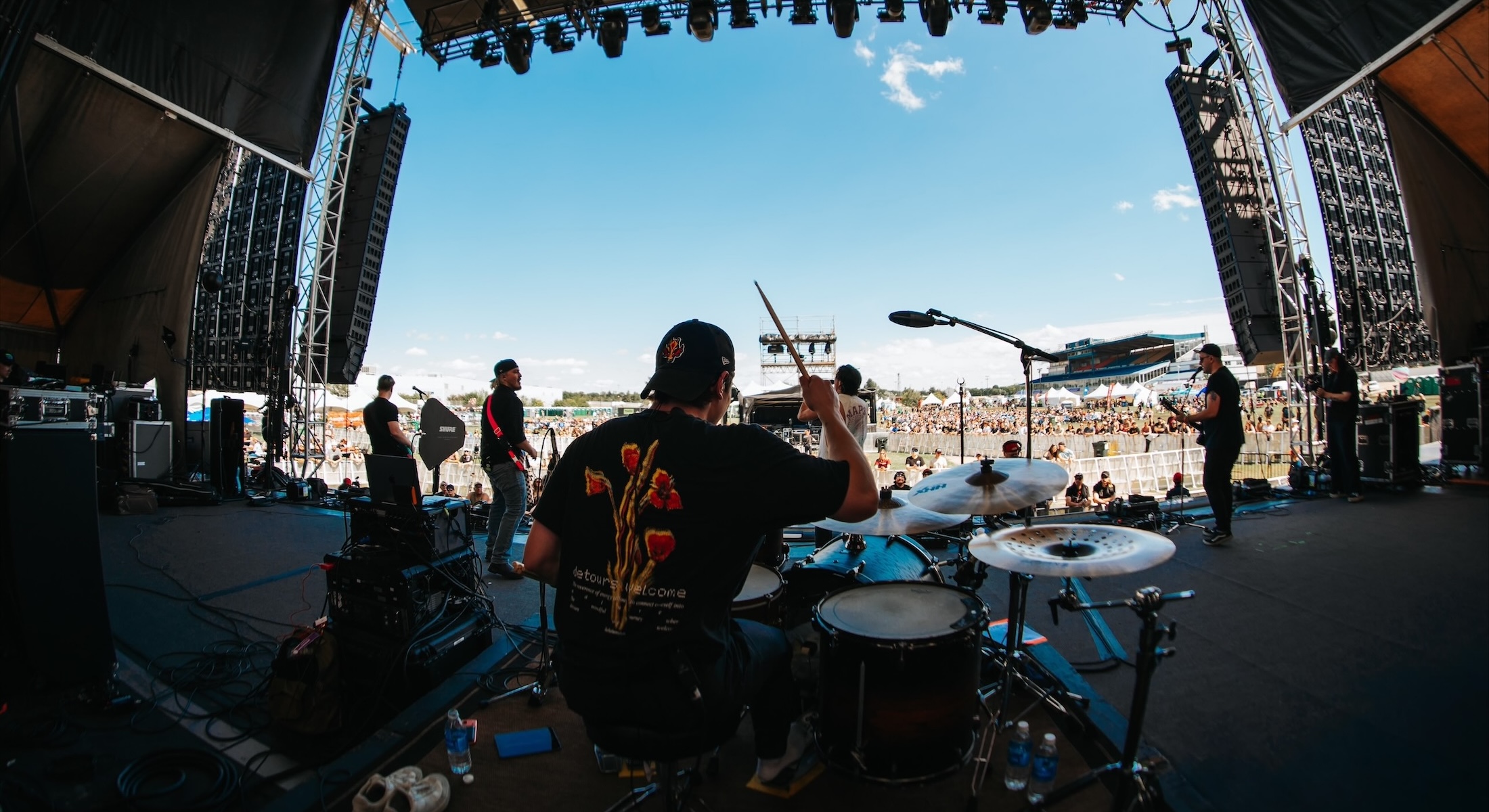 A rock band plays on a large outdoor stage at a music festival. The picture is taken from the back of the stage facing the audience. In the foreground is the band&rsquo;s drummer at his drum kit, his back turned to the camera.