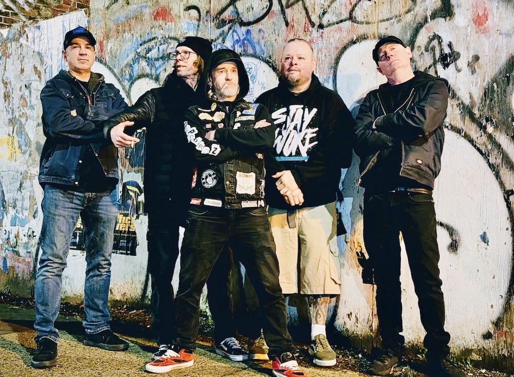 Five men pose for a photo in front of a white wall full of graffiti. They are wearing black jackets or hoodies, some of which are adorned with punk patches or graphics.