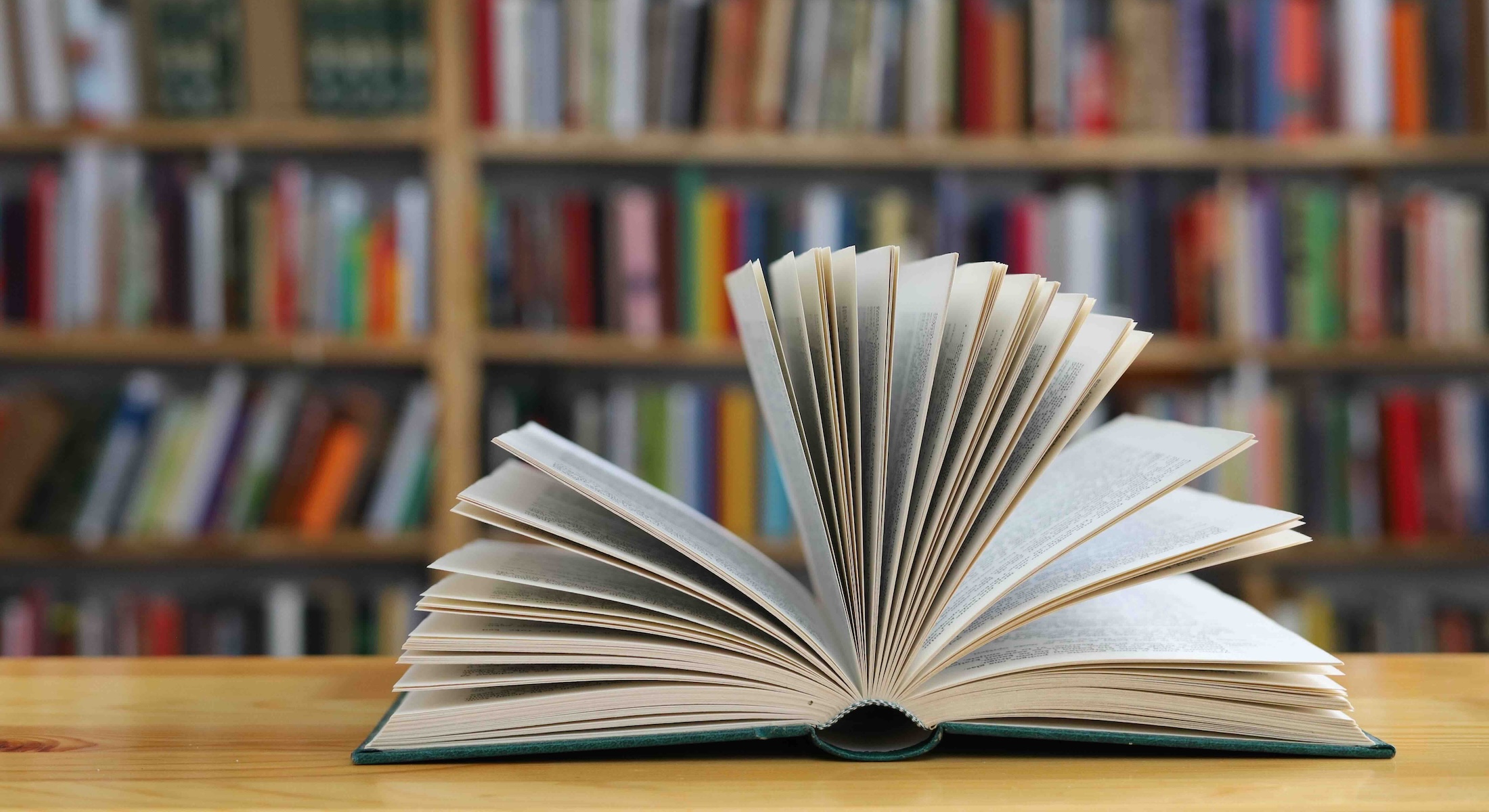 A book sits with its pages open on a wooden table in front of a large bookshelf.