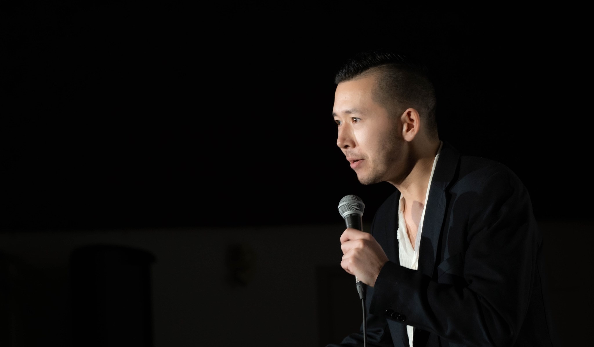 Trevor Jang speaks into a microphone on a darkened stage. He has short dark hair and is wearing a black blazer over a white top.