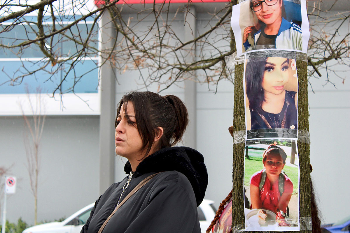 A woman with dark brown hair in a ponytail gazes into the distance. Behind her, posters show the faces of three young women and youth.