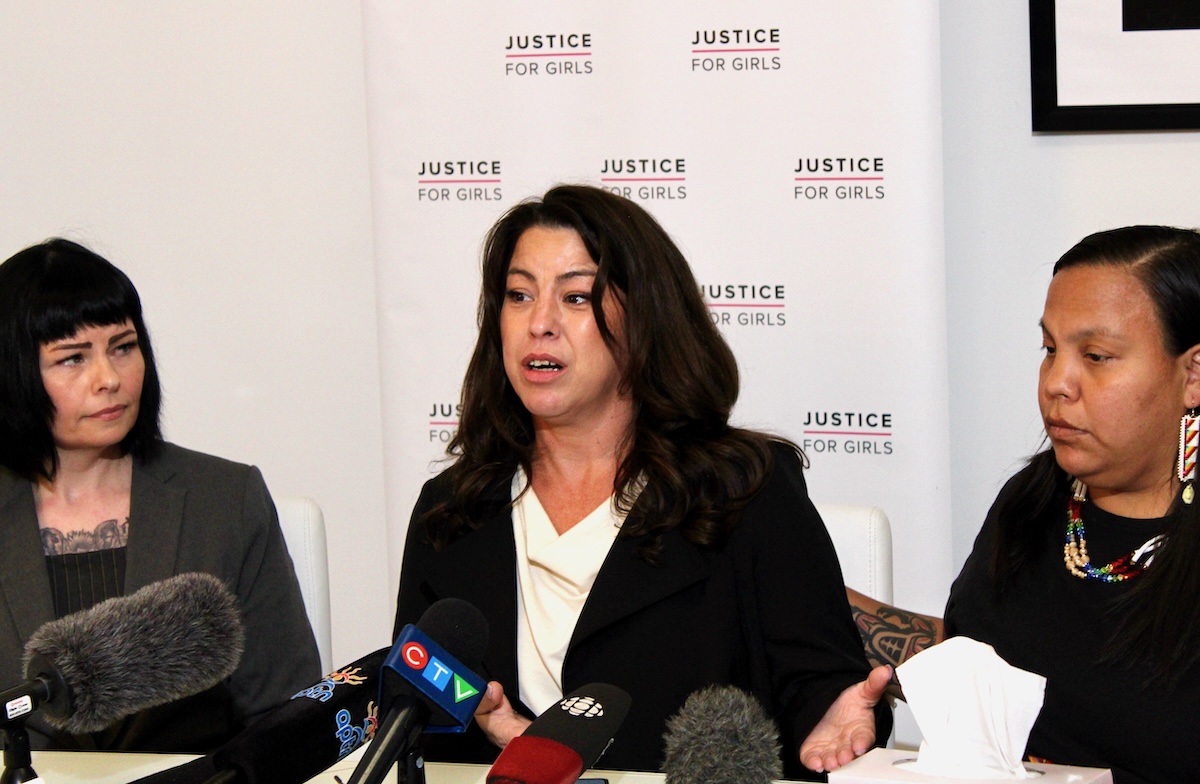 Three women with dark hair sit in front of a poster that reads &lsquo;Justice for Girls&rsquo; with several press microphones lined up in front of them.