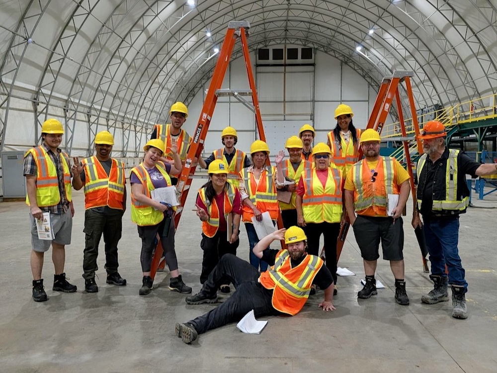 A large group of men and women pose wearing hard hats and high-vis vests inside a large industrial building.
