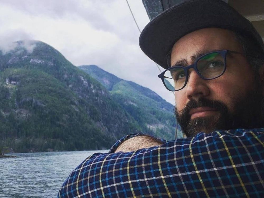 A light skin toned and bearded man wearing a baseball hat and a striped shirt looks on as a background of sea and hillside rolls by behind him.