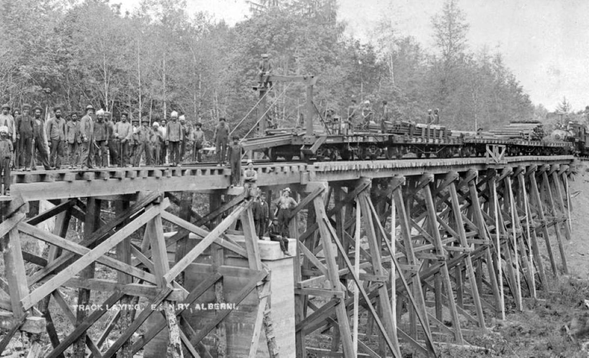 Archive black and white photo show many labourers standing and working on an elevated railway track, with a long train loaded with timber behind them.