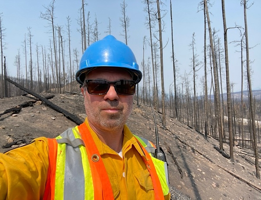A man wearing a high-vis vest, blue hard hat and sunglasses stands in front of a burned forest. 