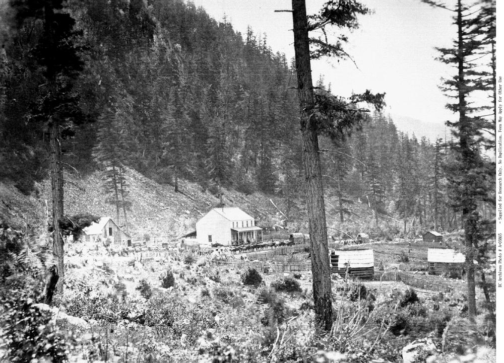 A black and white photo from long ago. A farmhouse-looking building sits in front of a hillside. A horse-drawn wagon train appears to be parked in front of it.