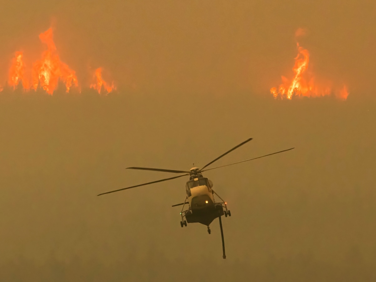 A helicopter flies away from two massive banks of flames.