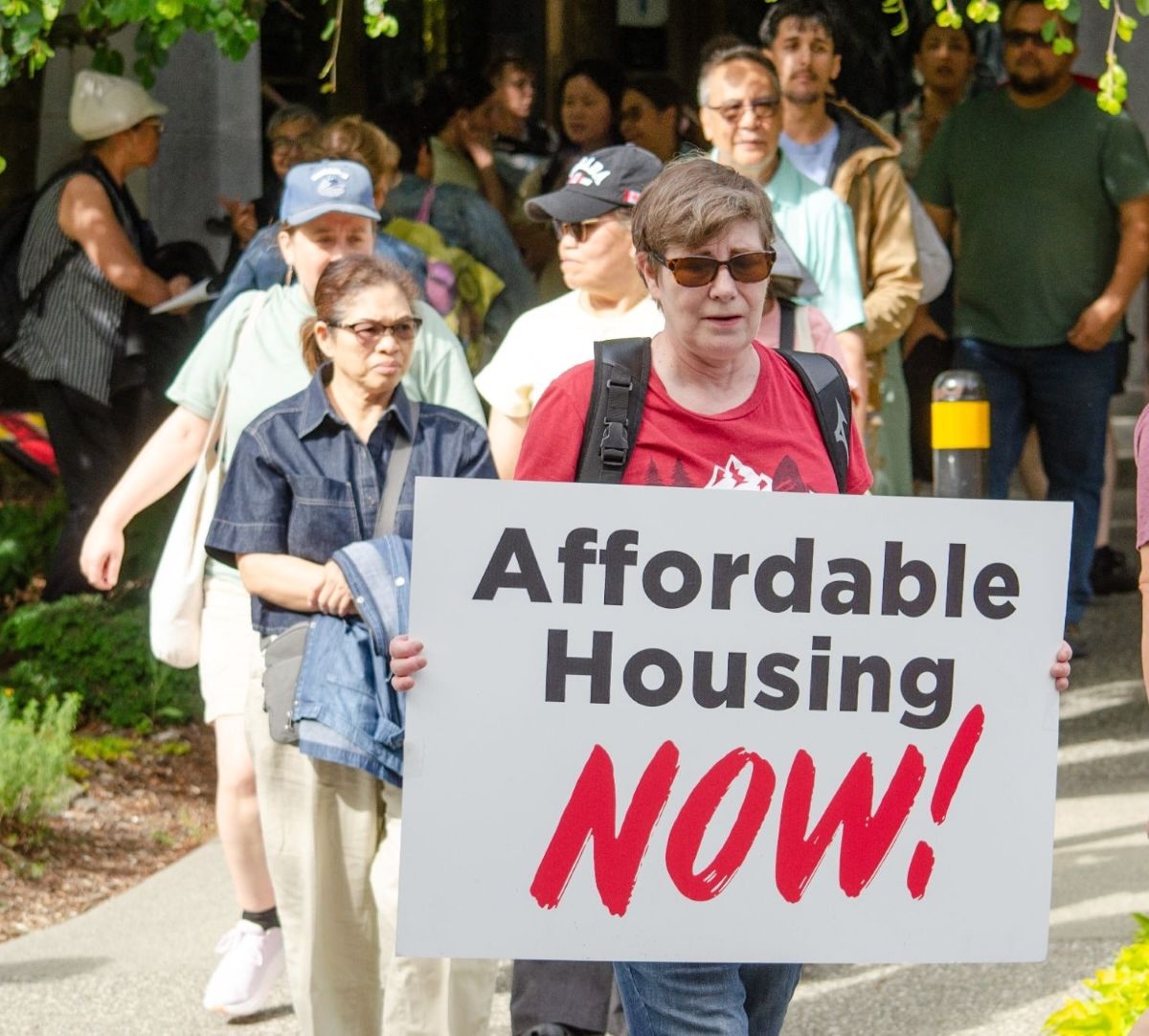 A middle-aged woman with short brown hair holds a sign that says, &lsquo;Affordable housing now!&rsquo;