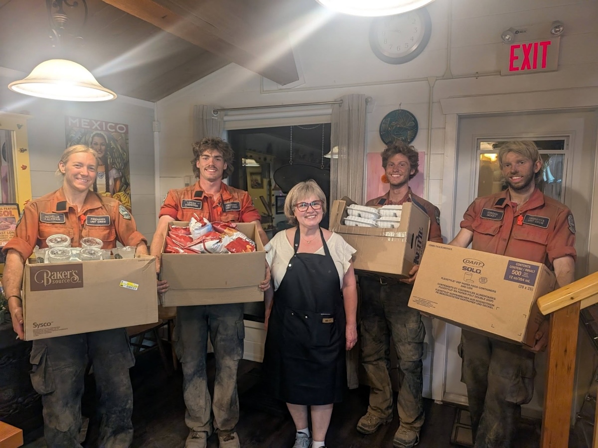 A woman in a smock stands flanked by four firefighters wearing orange shirts and with faces suggesting they came straight from the fire line. They&rsquo;re holding large cardboard boxes full of food.