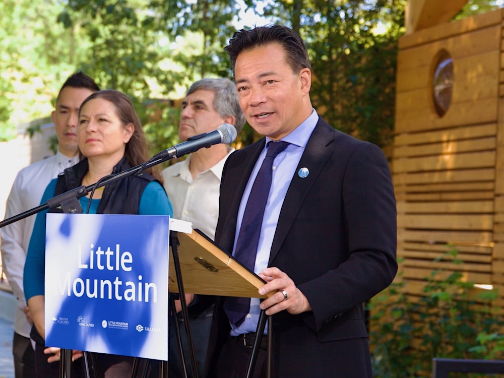 A middle-aged man with short black hair wearing a blue suit speaks at a lectern. 