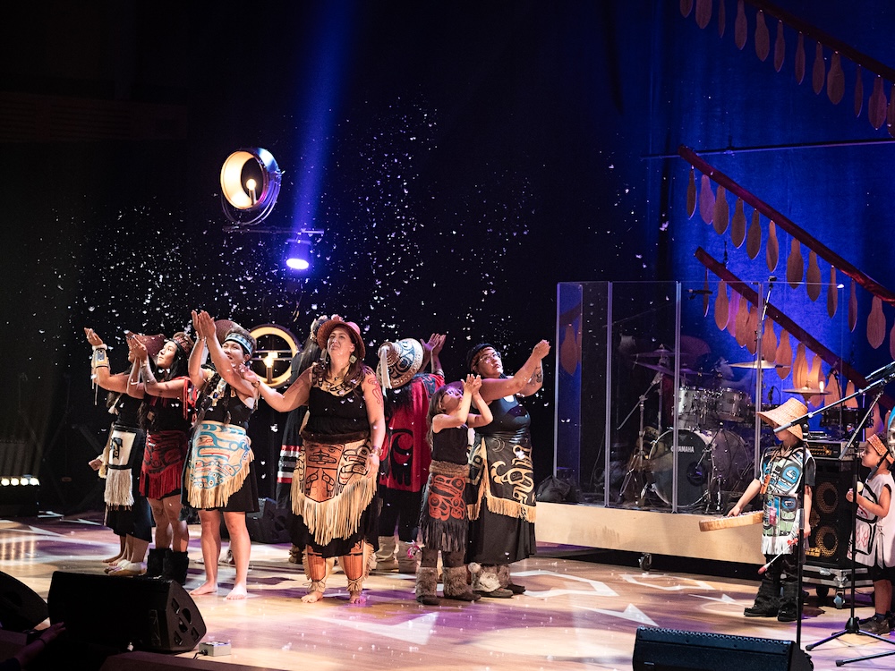 A group of performers gather onstage at the 2025 Come Toward the Fire festival. They stand in a circle with their backs facing each other and throw confetti in the air.