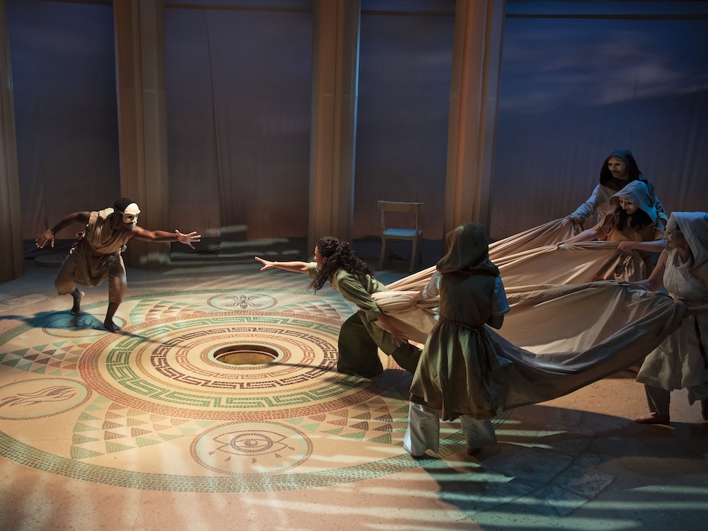 Polynices reaches for Antigone's outstretched hand. Antigone is held back by a length of drapery held by the masked Chorus.