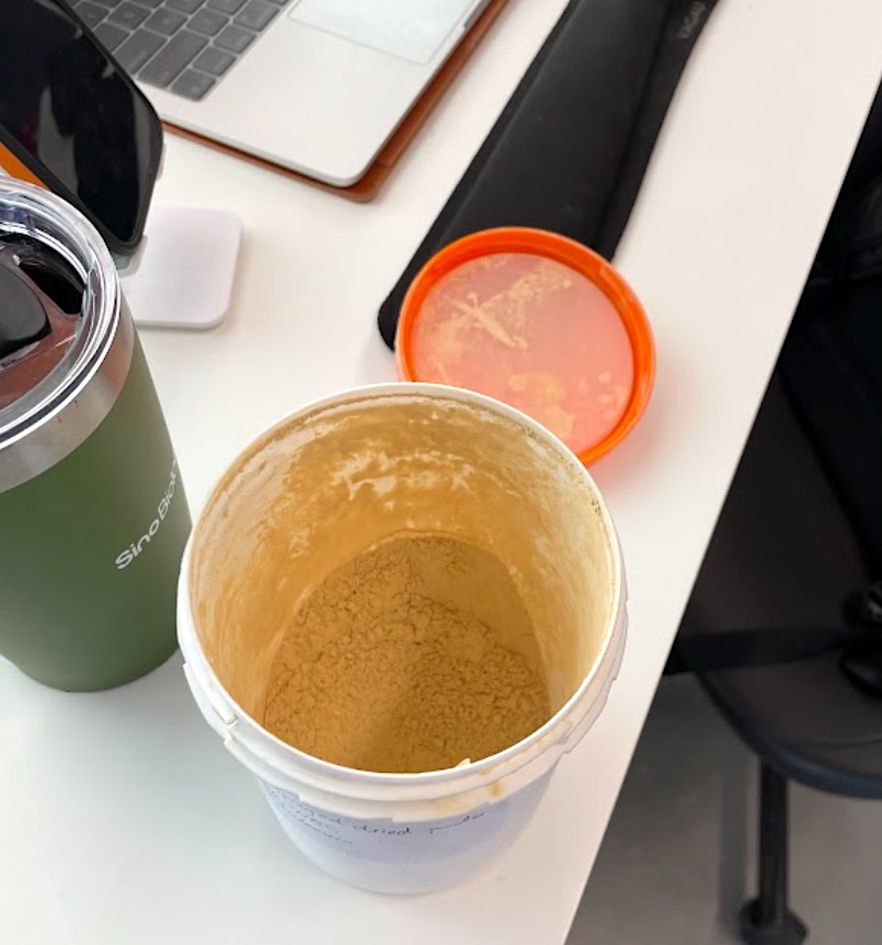 A white tub with the lid off on a desk, revealing an orange-brownish powder. There is a laptop, thermos and iPhone in the background. 