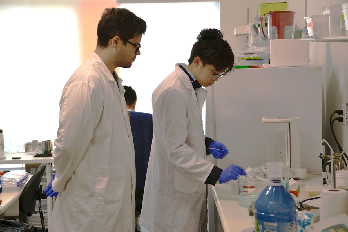 A pair of scientists in white lab coats run tests in an office. A man on the left observes, while a man on the right conducts the test in front of a machine. 
