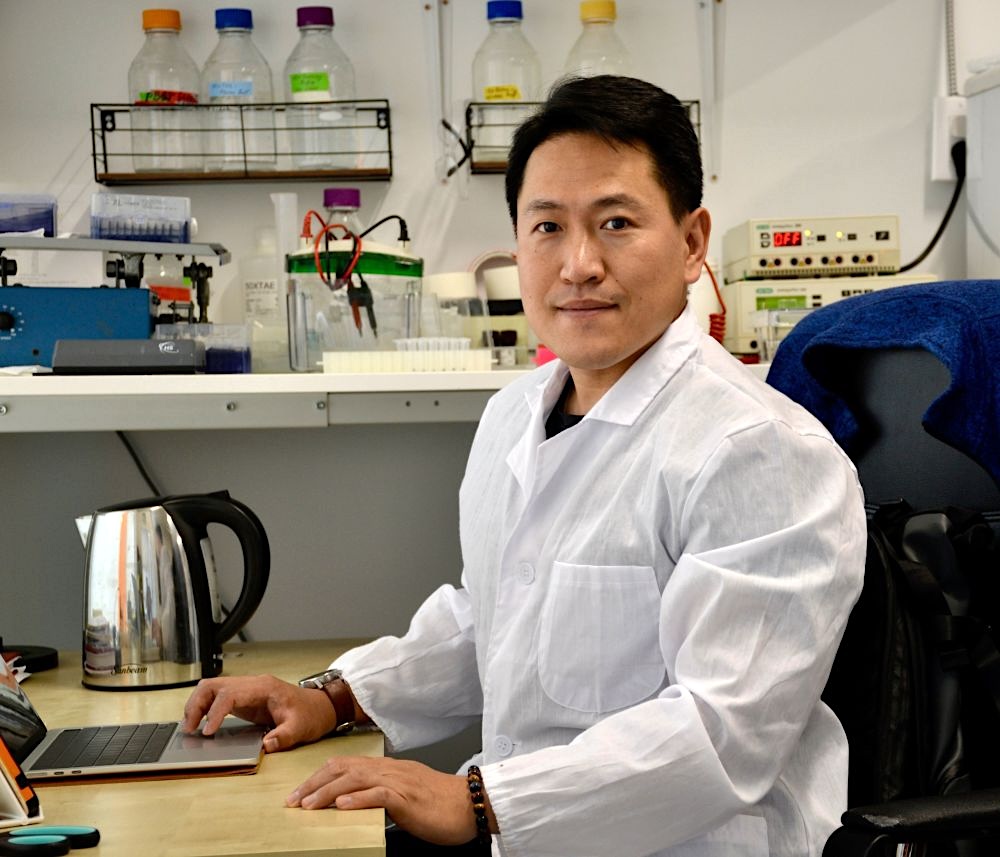 A man, Mingyang Sun, in a white lab coat stares and smiles directly into the camera. There&rsquo;s science equipment in the background and a kettle on the desk. 